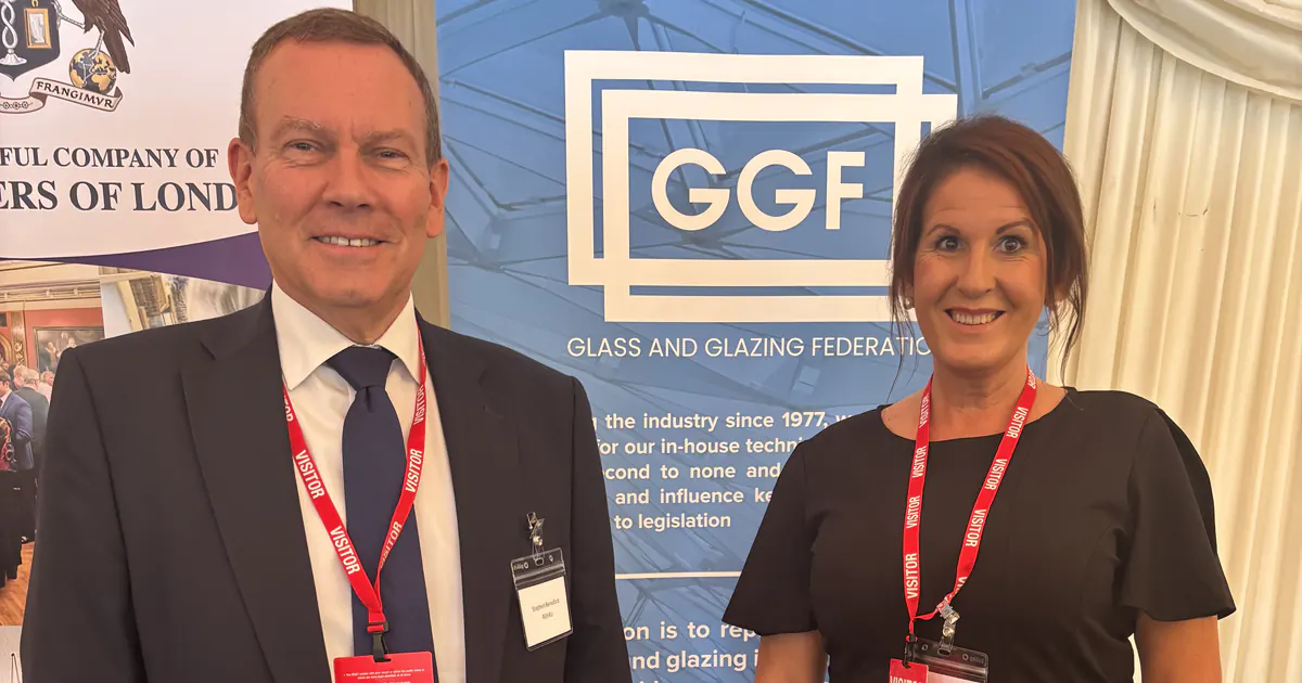 Two people standing indoors in front of a Glass and Glazing Federation banner, both wearing visitor badges
